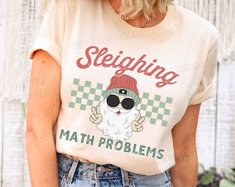 May include: A cream-colored t-shirt with the words "Sleighing MATH PROBLEMS" in red and green. The graphic features a cartoon Santa Claus wearing sunglasses and a beanie, giving a peace sign. A green and white checkered pattern is behind the Santa Claus graphic.