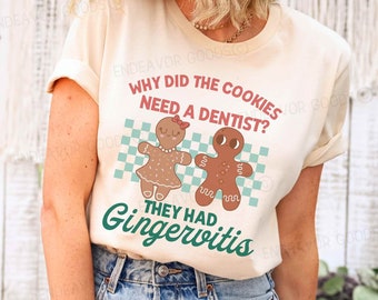 May include: A cream-coloured t-shirt with a graphic featuring two gingerbread people and the text "WHY DID THE COOKIES NEED A DENTIST? THEY HAD GINGERWITIS." The design includes a checkered background in light blue and white.