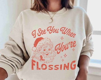 May include: A sand-colored sweatshirt with the text "I See You When You're Flossing" in red, along with a Santa Claus graphic. The design includes stars and is a festive holiday theme.