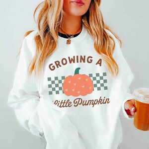 May include: White sweatshirt with the text "GROWING A little Pumpkin" in brown script. The design includes a peach-colored pumpkin with heart details and a green stem, set against a checkered background. The word "white" is at the bottom.