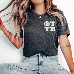 May include: A dark gray t-shirt with a white graphic of a horseshoe and the letters "OTB".