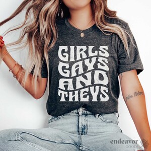 May include: A dark gray t-shirt with a white graphic that says "Girls, Gays, and Theys" in a retro font.