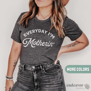 Op de afbeelding: Donkergrijs T-shirt met de witte tekst "EVERYDAY I'M Motherin'". Het T-shirt met ronde hals wordt gedragen met een donkerblauwe jeans. De afbeelding toont ook een hoed en sieraden.