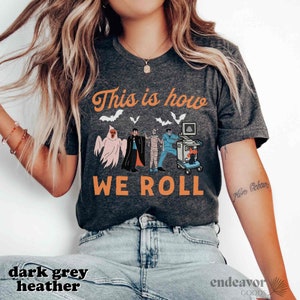 May include: Dark grey heather t-shirt with the text "This is how we roll" in orange. The shirt features a Halloween-themed graphic with a ghost, vampire, mummy, and medical equipment.