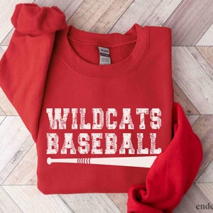 May include: A red sweatshirt with the words "WILDCATS BASEBALL" in white, distressed lettering. A white baseball bat graphic is below the text. The word "red" is in the bottom left corner.