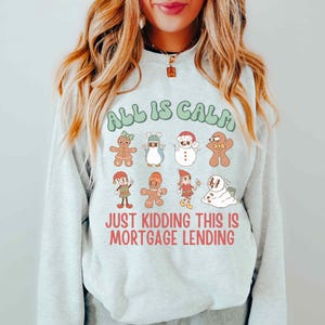 May include: Ash-colored sweatshirt with the text "ALL IS CALM" above a series of cartoon holiday characters. Below, the text reads "JUST KIDDING THIS IS MORTGAGE LENDING". The sweatshirt has a crew neck and long sleeves.