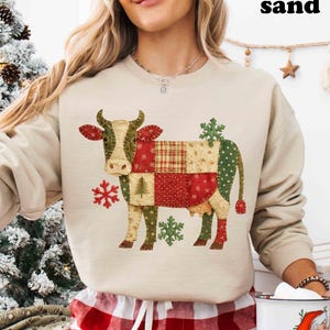 May include: A sand-colored sweatshirt featuring a patchwork cow design with red, green, and beige patterns. The cow has snowflake accents. The model is wearing red and white plaid pajama pants and holding a mug with a Santa Claus graphic.
