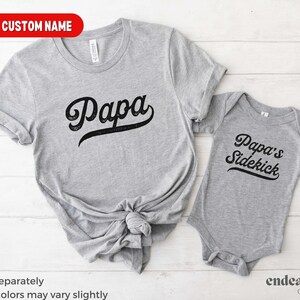 May include: Two grey t-shirts with black text. The larger shirt says "Papa" in a vintage baseball font. The smaller shirt says "Papa's Sidekick" in the same font.