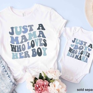 May include: A white t-shirt with the text "Just a Mama who loves her boy" in blue and gray letters. A white baby onesie with the text "Just a Boy who loves his Mama" in blue and gray letters. Both shirts have a heart design.