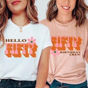 May include: Two white t-shirts with a retro-style graphic design. The first shirt says "Hello Fifty" with a pink flower. The second shirt says "Fifty Birthday Crew" with a pink flower.