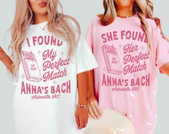 May include: Two white t-shirts with pink text. The text on the left shirt reads "I found my perfect match est. 2024 Anna's Bach Asheville, NC". The text on the right shirt reads "She found her perfect match est. 2024 Anna's Bach Asheville, NC".