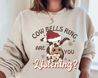 May include: Sand-colored sweatshirt with a graphic of a cow wearing a Santa hat. The text reads "Cow Bells Ring Are You Listening?" in a retro font, with snowflakes and stars. A festive holiday design.
