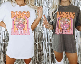 May include: Two oversized t-shirts with a retro 70s design featuring a disco ball and flowers. The white shirt says "Disco Bride" and the gray shirt says "Dancing Queen".