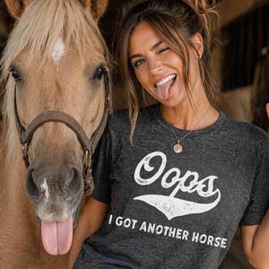 May include: Dark grey heather t-shirt with the text "Oops I Got Another Horse" in white script. The image also features a horse and a person.