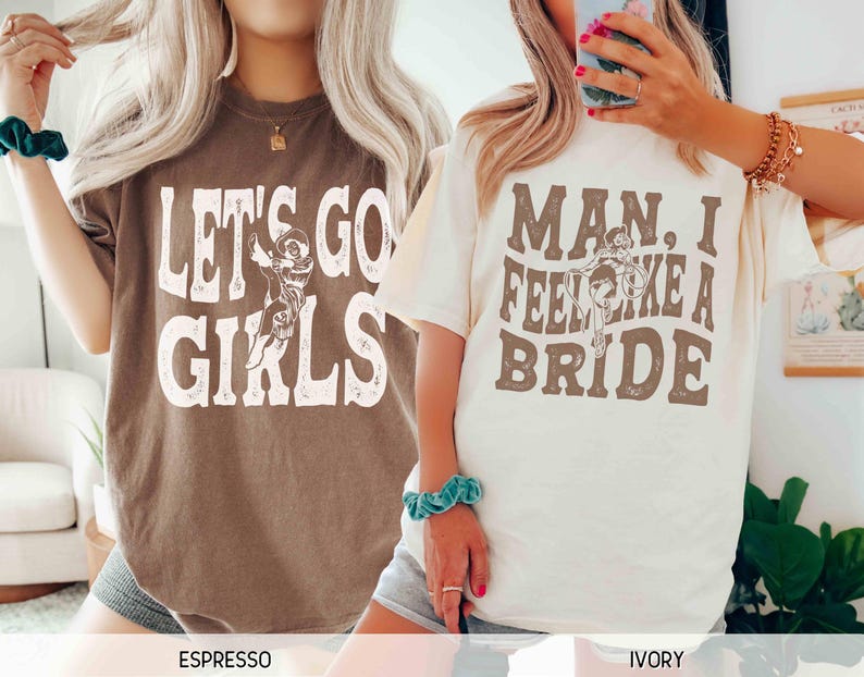 K&ouml;nnte beinhalten: Zwei T-Shirts, eines in Espresso und eines in Elfenbein, jeweils mit einem Grafikdesign. Das Espresso-Shirt zeigt "LET'S GO GIRLS" in Wei&szlig;, und das elfenbeinfarbene Shirt zeigt "MAN, I FEEL LIKE A BRIDE" in Braun. Beide haben einen Vintage-Stil.