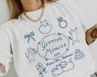 Può includere: Felpa bianca con un design grafico blu con gatti, un anello, calici di champagne e la scritta "Getting Meowied 2026 Future Mrs. Robinson." La parola "white" è stampata in basso.