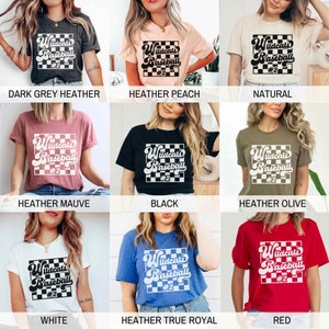 May include: A collection of t-shirts in various colors, each featuring the text "Wildcats Baseball #2" in a retro font within a checkered square. Colors include dark grey, peach, natural, mauve, black, olive, white, blue, and red.