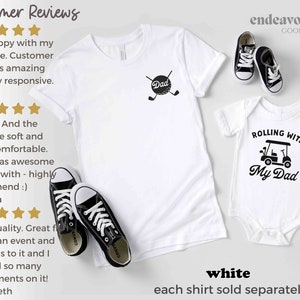 May include: Two white t-shirts with a golf theme. The larger shirt says "Dad" with a golf ball and clubs. The smaller shirt says "Rolling with My Dad" with a golf cart graphic. Both shirts are on a white background with black text and graphics.