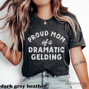 以下が含まれることがあります： 白い文字で「Proud Mom of a Dramatic Gelding」とプリントされた、濃いグレーのヘザーTシャツです。