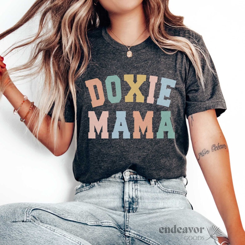 May include: Dark gray t-shirt with the text "DOXIE MAMA" in colorful block letters. The shirt is short-sleeved and has a crew neck. The model is wearing light blue jeans.