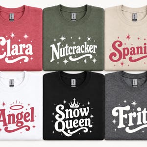 May include: Six crewneck sweatshirts in red, olive green, beige, white, black, and dark gray. Each features a unique word or phrase in a retro font with star accents: Clara, Nutcracker, Spanish, Angel with a halo, Snow Queen with a crown, and Fritz.