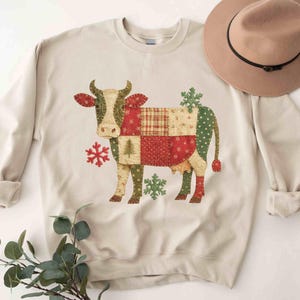 May include: A cream-colored sweatshirt featuring a patchwork cow design with red, green, and beige patterns. The cow has snowflake accents. A brown hat with a black band rests on the sweatshirt.