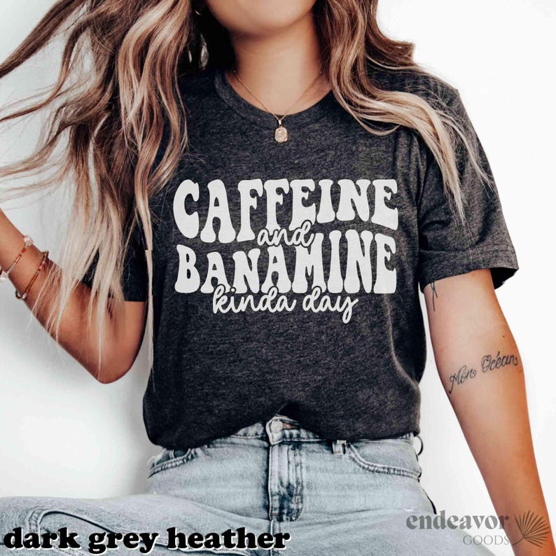 May include: Dark grey heather t-shirt featuring the white text "Caffeine and Bananine kinda day" in a retro font. The top is a short-sleeved crew neck.