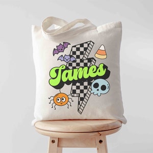 May include: A cream-colored tote bag with a Halloween-themed design. The design includes the name "James" in green and black, a checkered lightning bolt, bats, a spider, a skull, and candy corn.