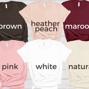 May include: Six different colored t-shirts laid out on a wooden surface. The colors are brown, heather peach, maroon, pink, white, and natural.