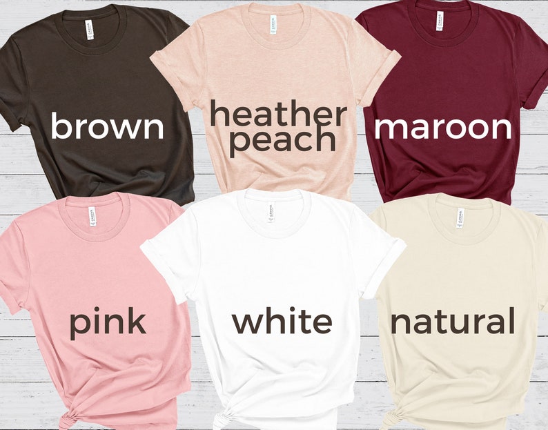 May include: Six different colored t-shirts laid out on a wooden surface. The colors are brown, heather peach, maroon, pink, white, and natural.