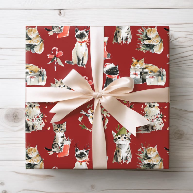 May include: A wrapped gift with red wrapping paper featuring a pattern of watercolor cats in festive attire. The gift is tied with a cream-colored satin ribbon and bow, set against a white wooden background.