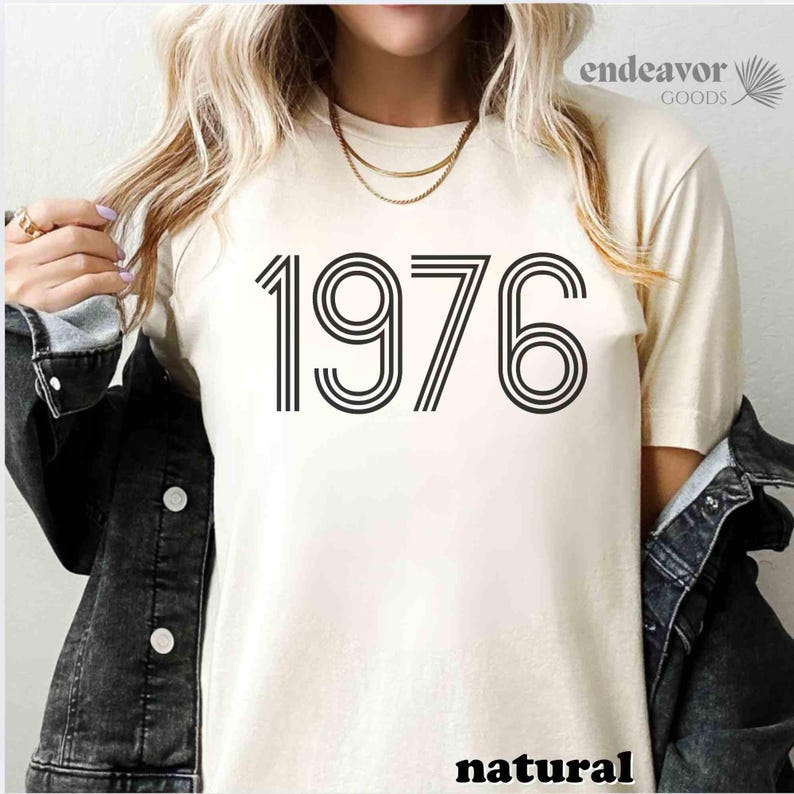May include: Cream-coloured t-shirt featuring the year "1976" in black, outlined block letters. The word "natural" is printed at the bottom. The shirt is part of a clothing collection.