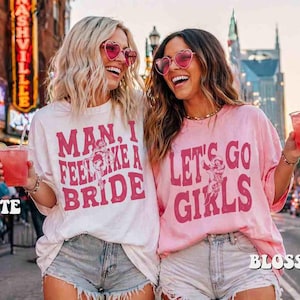 K&ouml;nnte beinhalten: Zwei Frauen tragen herzf&ouml;rmige Sonnenbrillen und T-Shirts mit den Aufschriften "Man, I Feel Like a Bride" und "Let's Go Girls". Sie halten rosa Getr&auml;nke mit Strohhalmen. Die Shirts sind wei&szlig; und rosa.