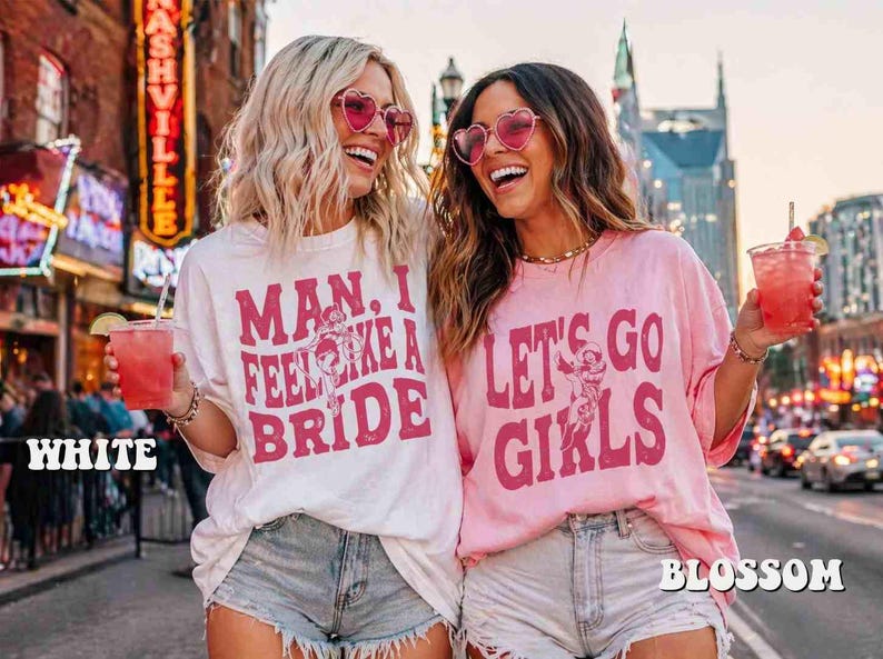 K&ouml;nnte beinhalten: Zwei Frauen tragen herzf&ouml;rmige Sonnenbrillen und T-Shirts mit den Aufschriften "Man, I Feel Like a Bride" und "Let's Go Girls". Sie halten rosa Getr&auml;nke mit Strohhalmen. Die Shirts sind wei&szlig; und rosa.