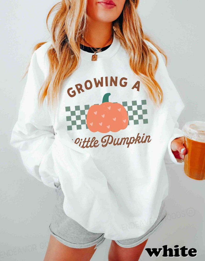 May include: White sweatshirt with the text "GROWING A little Pumpkin" in brown script. The design includes a peach-colored pumpkin with heart details and a green stem, set against a checkered background. The word "white" is at the bottom.