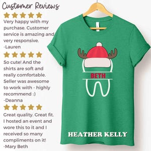以下が含まれることがあります： 赤と白の帽子と角、名前「BETH」、名前「HEATHER KELLY」の白い文字が特徴の、お祝いのデザインの緑色のTシャツ。木製のハンガーにシャツが表示されています。