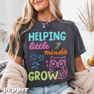 May include: Dark gray t-shirt with the text "HELPING little minds GROW" in various colors. The design includes stars and an owl with books. The shirt is a comfortable, casual style.