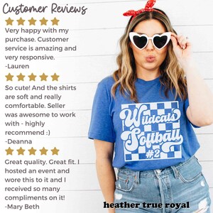 May include: A blue t-shirt with a white checkered design and the text "Wildcats Softball #2" in a retro font. The shirt is being worn by a person with long blonde hair and heart-shaped sunglasses.