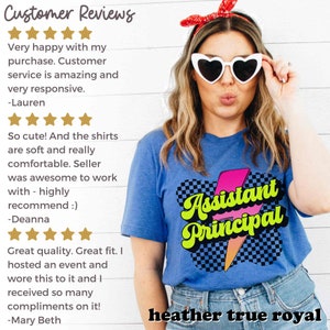 K&ouml;nnte beinhalten: Ein blaues T-Shirt mit einem pinkfarbenen und gelben Blitz-Motiv, auf dem in einer Retro-Schrift "Assistant Principal" steht. Das Shirt wird von einer Person mit blonden Haaren und herzf&ouml;rmigen Sonnenbrillen getragen. Der Text "heather true royal" ist am unteren Rand des Bildes sichtbar.