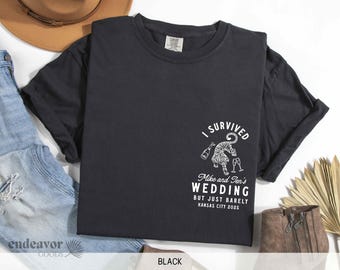 May include: Black t-shirt with a white graphic of a tiger holding a glass of champagne. The text reads "I Survived Mike and Jen's Wedding But Just Barely Kansas City 2025".