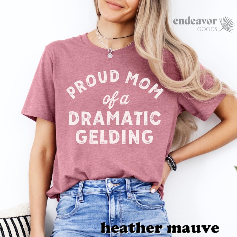 以下が含まれることがあります： ヘザーモーヴのTシャツに、白い文字で「Proud Mom of a Dramatic Gelding」と書かれています。