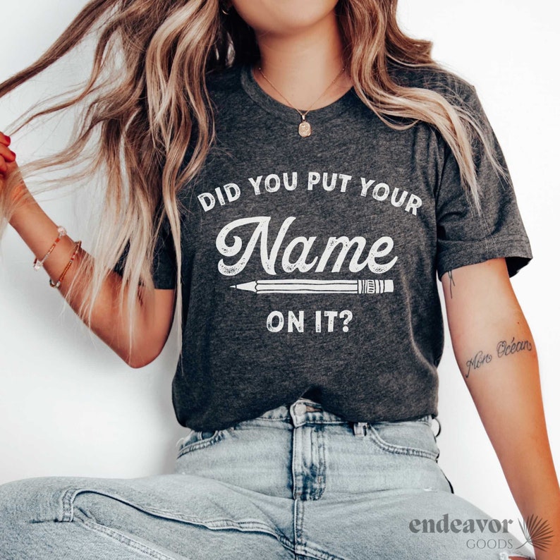May include: A dark gray t-shirt with the text "Did you put your name on it?" in white. A white pencil graphic is below the text.