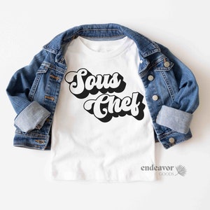 K&ouml;nnte beinhalten: Wei&szlig;es T-Shirt mit schwarzem Retro-Text, der "Sous Chef" lautet. Das Shirt ist gefaltet und auf einer blauen Jeansjacke abgelegt.