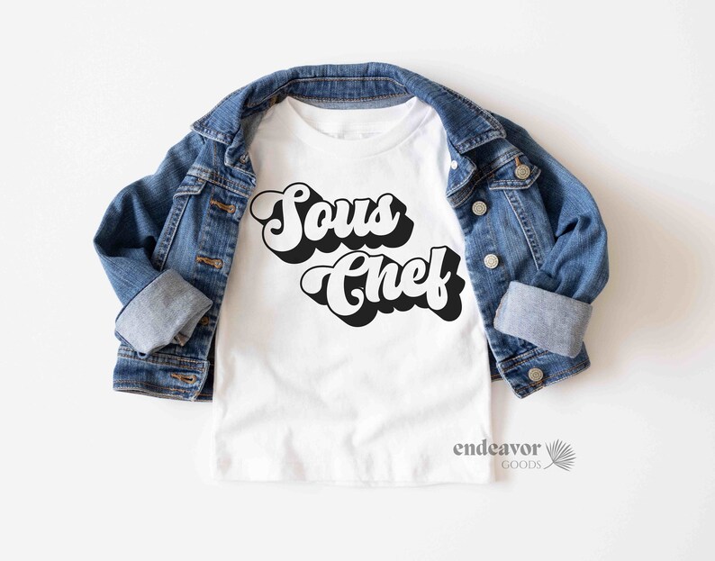 K&ouml;nnte beinhalten: Wei&szlig;es T-Shirt mit schwarzem Retro-Text, der "Sous Chef" lautet. Das Shirt ist gefaltet und auf einer blauen Jeansjacke abgelegt.