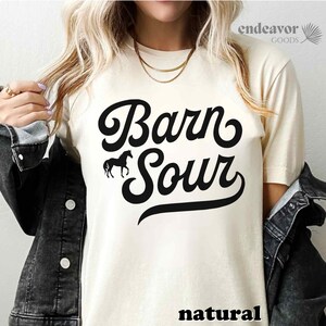 May include: A white t-shirt with a black graphic that says "Barn Sour" in a retro font. A small silhouette of a horse is to the left of the text. The word "natural" is printed in black at the bottom of the shirt.