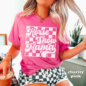 K&ouml;nnte beinhalten: Ein pinkfarbenes T-Shirt mit einem wei&szlig;en karierten Hintergrund und dem Text "Horse Show Mama" in einer Retro-Schrift.
