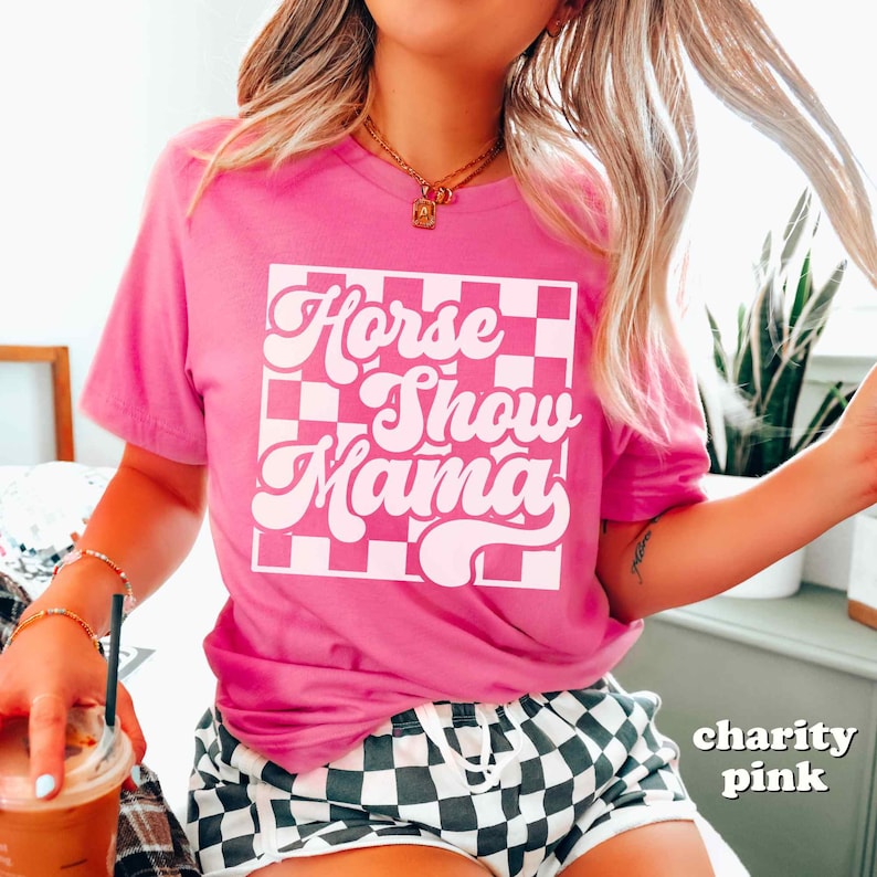K&ouml;nnte beinhalten: Ein pinkfarbenes T-Shirt mit einem wei&szlig;en karierten Hintergrund und dem Text "Horse Show Mama" in einer Retro-Schrift.