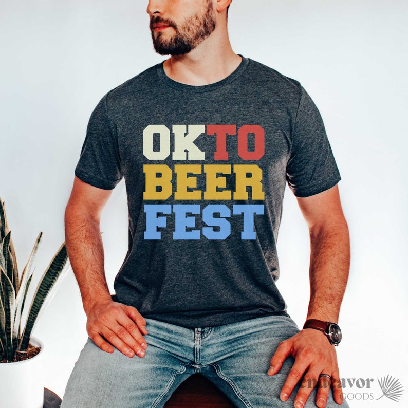 Op de afbeelding: Donkergrijs t-shirt met de tekst "OK TO BEER FEST" in een retro lettertype. De tekst is gelaagd met witte, rode, gele en blauwe kleuren. Het shirt heeft korte mouwen en is gemaakt van een zacht materiaal.