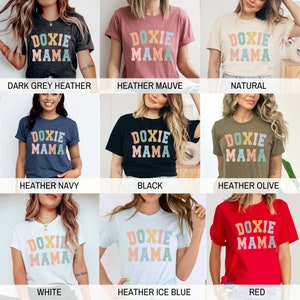 May include: A collection of t-shirts in various colors, each featuring the text "DOXIE MAMA" in a colorful, retro font. The shirts are displayed on models, showcasing the different color options available, including dark grey, mauve, natural, navy, black, olive, white, ice blue, and red.