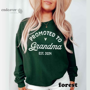 May include: A dark green sweatshirt with the text "Promoted to Grandma Est. 2024" in white lettering. The text is in a circle with a small heart above the word "Grandma".